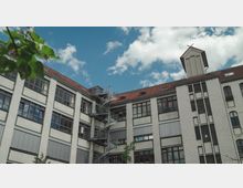 Backsteinbau mit weißen Klinkern und roten Dächern, großen Fenstern und einer Wendeltreppe an der Außenfassade, unter einem blauen Himmel mit wenigen Wolken. Im Vordergrund sind grüne Blätter und Bäume sichtbar.