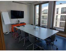 Konferenzraum mit großem rechteckigem Tisch, umgeben von Stühlen auf Rollen. Im Hintergrund steht ein Flipchart sowie ein orangefarbener Schrank mit einem Wandbildschirm darüber. Große Fenster bieten Blick auf moderne Gebäude und einen Balkon.
