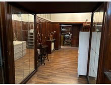 Innenansicht eines holzvertäfelten Raums mit Parkettboden, großen Glasvitrinen und Schrankelementen an den Wänden. Im Raum stehen Arbeitsmöbel und Geräte wie ein kleiner Tisch, Schubladenschränke und eine Leiter; im Hintergrund führt eine Öffnung in einen weiteren Raum mit Regalen.