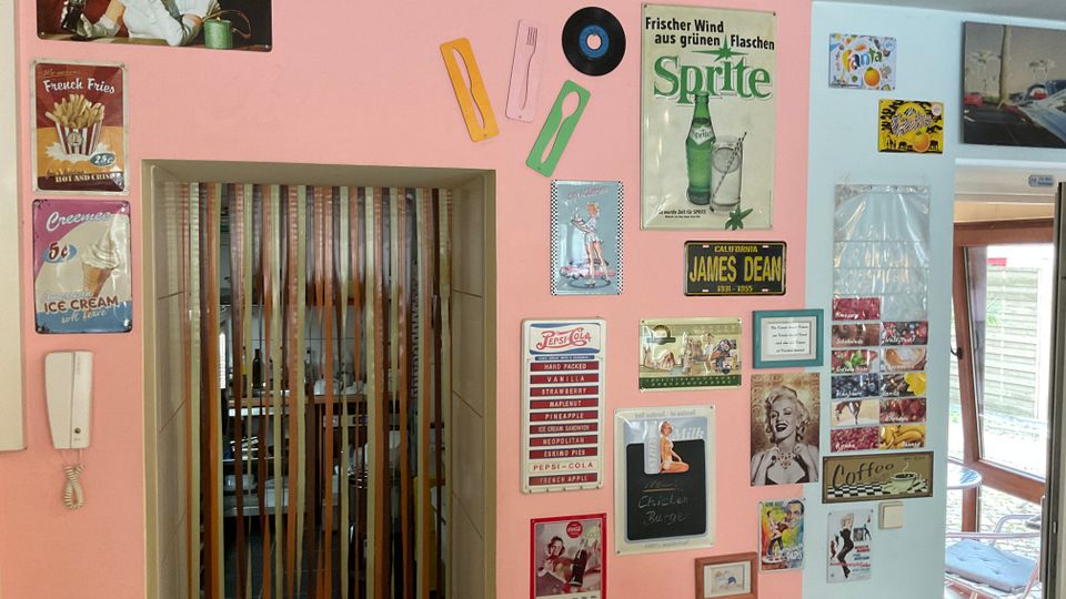 Innenraum mit rosa und hellblauen Wänden, dekoriert mit zahlreichen Retro-Blechschildern und Plaketten (u. a. Getränke- und Diner-Motive). In der Mitte befindet sich eine Türöffnung mit Lamellenvorhang, darunter ein Heizkörper; rechts führt ein Glasdurchgang zu einem hellen Wintergartenbereich.