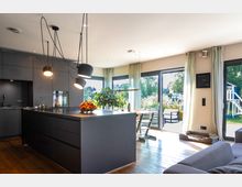 Modernes Wohn- und Esszimmer mit offener Küche, ausgestattet mit dunklen Einbauschränken, einer großen Kücheninsel und Hängelampen. Große Fenster und Glastüren führen zu einem Garten, wodurch viel Tageslicht in den Raum fällt, ergänzt durch einen Holzboden und helle Vorhänge.