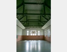 Innenansicht einer Turnhalle mit hoher, gewölbter Decke und sichtbaren Holzbalken. Der Raum hat helle Wände, große Fenster, die Tageslicht hereinlassen, sowie einen Basketballkorb an der Stirnwand über einem hellen, glatten Boden.