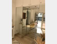 Eingangsbereich mit einem großen Spiegelschrank, weißen Oberschränken, einer hellen Fliesengestaltung und Teppichen. Durch eine Glastür fällt Tageslicht in den Raum, und ein Teil des Außenbereichs ist sichtbar.