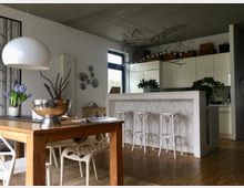 Offene Küche und Essbereich mit modernen und natürlichen Elementen. Ein Holztisch mit weißen Stühlen steht im Vordergrund, während im Hintergrund eine Kücheninsel mit weißer Marmoroptik und Barhockern sowie ein Schrank mit Pflanzen und Körben zu sehen sind.