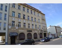 Straßenszene mit einem historischen, vierstöckigen Gebäude in gelber Fassade mit weißen Verzierungen und großen Fenstern. Im Erdgeschoss befindet sich eine Gaststätte mit Vordach und weiteren geschlossenen Schaufenstern; mehrere parkende Autos stehen entlang der Straße.