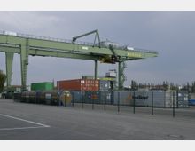 Ein Güterbahnhof oder eine Hafenanlage mit einem großen grünen Portalkran der Marke "BEHALA", gestapelten Frachtcontainern in verschiedenen Farben und eingezäuntem Areal. Im Hintergrund sind Bäume und vereinzelt Gebäude erkennbar.