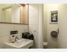 Modernes Badezimmer mit weiß gefliesten Wänden, einem Waschbecken mit Spiegel und Holzrahmen sowie einem Handtrockner von Dyson. Im Hintergrund ist eine teilweise geöffnete Tür, die zu einer Toilette führt, sowie ein Wandbild mit einer Werbegrafik zu sehen.