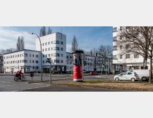 Straßenszene mit mehreren weißen, mehrstöckigen Wohn- und Geschäftshäusern im minimalistischen Stil der Moderne. Im Vordergrund stehen Ampeln, parkende Autos, Radfahrer, sowie eine Litfaßsäule mit Werbung, umgeben von Bäumen und einer Kreuzung.