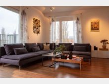 Modernes Wohnzimmer mit dunklem Ecksofa, Holzboden und großen Fenstern mit transparenten Vorhängen. Ein Couchtisch mit Dekorationen und Pflanzen steht im Zentrum, während die Wände mit Kunst und einem gerahmten Poster geschmückt sind.