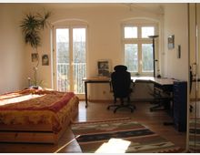 Helles Schlafzimmer und Arbeitszimmer kombiniert, mit Holzboden und großen Fenstern. Ein Bett mit bunter Decke steht links, ein Schreibtisch mit Schreibtischstuhl sowie Regalen rechts; an der Wand hängt ein großer Spiegel.