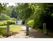 Ein Weg in einem grünen Park führt zu einem Geländer am Ufer eines Flusses, im Hintergrund sind dichte Bäume und ein Steg zu sehen. Drei betonierte Poller mit Graffiti markieren den Wegesrand, während eine Person am Geländer steht und auf das Wasser blickt.