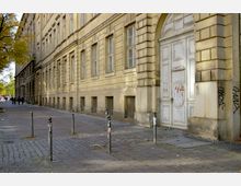 Historisches Gebäude mit heller Fassade und großen rechteckigen Fenstern; im Vordergrund befinden sich ein Pflasterweg, Poller und ein alter, bemalter Holztürbogen. Links führt ein Gehweg mit Bäumen und Fußgängern entlang der Straße.