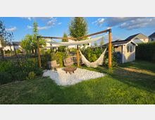 Garten-Lounge-Bereich mit Holzpergola, darüber einem Sonnensegel und seitlich einer Hängematte, auf einer Fläche aus weißen Kieseln mit Feuerstelle und Hängesesseln. Im Hintergrund sind Blumenbeete, Rasen, ein kleines Gartenhaus und Wohnhäuser zu sehen.