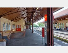Ein überdachter S-Bahnhof mit Holzdach und Metallträgern, einer gelben Hauswand mit roten Türen und gewölbten Fenstern. Im Hintergrund befinden sich Gleise, Sitzbereiche, Ticketautomaten und ein SOS-Säule im Vordergrund.