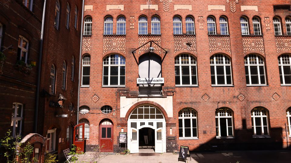 Innenhof mit mehrstöckigem Backsteingebäude und dekorativen Ziegelmustern, großen rundbogigen und rechteckigen Fenstern sowie einem zentralen Rundbogen-Eingang. Über dem Eingang hängt ein Schild „Sophiensäle“, davor liegt eine gepflasterte Fläche mit wenigen Pflanzen und Aufstellschildern.