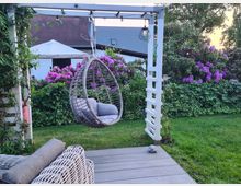 Garten mit einer Terrasse aus Holzplanken, einem Hängesessel aus Rattan und einem weißen Pergola-Gerüst. Im Hintergrund eine grüne Hecke, blühende lila Rhododendren und ein Haus mit hellen Wänden und einem grauen Dach.