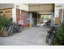 Vor einem Wohngebäude mit braunen Kacheln und einer grauen Fassade stehen mehrere Fahrräder geparkt. Ein Metallzaun mit Tor führt zu einem Durchgang, und an der Wand sind Graffiti sowie ein Kaugummiautomat sichtbar.