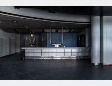 Leere Bar in einem dunklen Innenraum mit einer Metalltheke, einer abgehängten Decke mit Gitterstruktur und schlichten Hängelampen. Die Wände sind hellblau, flankiert von runden Säulen, und der Boden besteht aus dunklem Material.