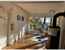 Wohnraum mit Holzboden und großen Glas-Schiebetüren, die auf eine Terrasse mit Blick auf Bäume und einen See führen. Im Vordergrund ein Kaminofen, im Hintergrund hängen Kleidungsstücke und Kindergegenstände, und ein Teppich liegt auf dem Boden.