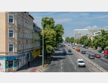 Straßenszene in einer Stadt mit mehrspuriger Straße, flankiert von mehrstöckigen Wohnhäusern auf der linken Seite und einem modernen Wohngebäude auf der rechten Seite. Entlang der Straße stehen Bäume, und Fahrzeuge sind in beide Richtungen unterwegs.