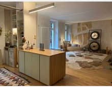 Wohnraum mit gemütlichem Ambiente, bestehend aus einem offenen Bereich mit einer Kücheninsel im Vordergrund, Regalen an einer Wand und einem Teppich mit Kissen und Decken im hinteren Bereich. Die Wand im Hintergrund ist mit abstrakter Kunst gestaltet, und zwei große Gongs stehen auf einem Ständer.