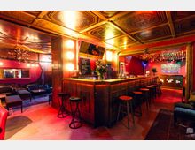 Innenansicht einer Bar mit langer Holztheke und einer Reihe von Barhockern, warm beleuchtet in Rot- und Goldtönen. Auffällig sind die dekorierte Kassettendecke, ein Loungebereich mit Sofas links und ein großer Durchgang zu einem hinteren Raum mit weiterer Theke und Beleuchtung.
