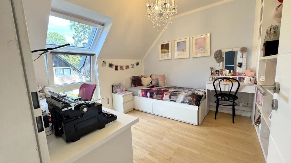 Helles Dachzimmer-Schlafzimmer mit schrägen Wänden, großem Dachfenster und Parkettboden. Zu sehen sind ein weißes Bett, ein Schminktisch mit Spiegel, Regale sowie ein Kronleuchter an der Decke.