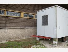 Ein kleiner, weißer Container mit Fenstergitter steht erhöht auf Betonblöcken neben einer Holzfassade mit langen, schmalen Fenstern, die von einem massiven Putzmauerwerk unterhalb der Holzverkleidung gestützt wird. Im Vordergrund befindet sich eine sandige Fläche mit etwas Grasbewuchs.