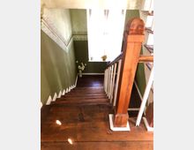 Holztreppe in einem Innenraum mit grün gestrichenen Wänden, verziert mit einer dekorativen Bordüre. Am Ende der Treppe steht ein Fenster mit durchscheinendem Licht, auf dessen Fensterbrett Pflanzen und Dekorationsgegenstände stehen.