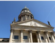 Neoklassizistisches Gebäude mit Säulen und einer verzierten Fassade, gekrönt von einer Kuppel mit einer goldenen Statue. Figuren und Reliefs schmücken den Giebel, während der Himmel klar und blau ist.