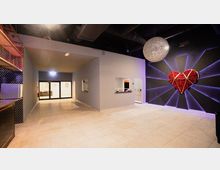 Ein moderner Innenraum mit hellen Fliesenboden, schwarzer Decke und sichtbaren Lüftungsrohren. An der rechten Wand befindet sich ein großes, dekoratives Herz aus roten Rosen, umgeben von violetter Beleuchtung, sowie eine glitzernde Kugel-Deckenleuchte.