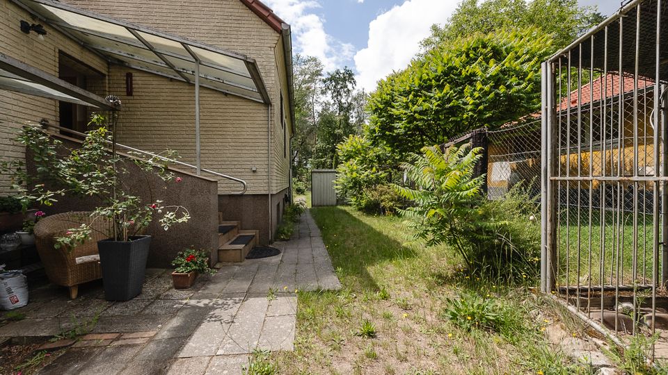 Schmaler Hof neben einem hellen Backsteinhaus mit Glasvordach über dem Eingang und einem gepflasterten Weg, der zu einem kleinen Schuppen am Ende führt. Rechts begrenzt ein Metallgitterzaun den Bereich, dahinter stehen dichte Sträucher und ein Nebengebäude mit rotem Ziegeldach.