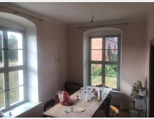 Ein einfacher Raum mit hellrosa Wänden und zwei großen, bogenförmigen Fenstern, die Tageslicht hereinlassen. In der Mitte steht ein Holztisch mit Stühlen, darauf befinden sich verschiedene Gegenstände wie Gläser und Küchengeräte; im Hintergrund ist eine Wiese sowie ein Backsteingebäude sichtbar.