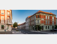 Straßenecke mit zwei mehrstöckigen Altbauten, die eine Fassade aus rot-braunem Ziegel und hellgrünen Stuckdetails aufweisen; daneben gepflegte Gehwege und eine ruhige Straße, gesäumt von weiteren historischen Gebäuden. Der Himmel ist klar und blau.