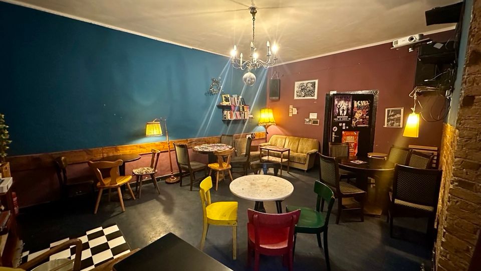 Gemütlicher Lounge- oder Caféraum mit blau- und bordeauxfarbenen Wänden, gemischten kleinen Tischen und Stühlen sowie Sofas und Stehlampen. An der Decke hängt ein Kronleuchter, an der Rückwand stehen Regale und gerahmte Bilder, daneben eine Tür mit Plakaten.