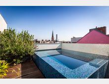 Rooftop-Terrasse mit einem kleinen Pool, der mit blau-mosaikfarbenen Fliesen verkleidet ist, umgeben von Pflanzen und hölzernem Bodenbelag. Im Hintergrund sind ein Kirchturm, ein moderner Hochhausturm und Dächer der Stadt zu sehen.