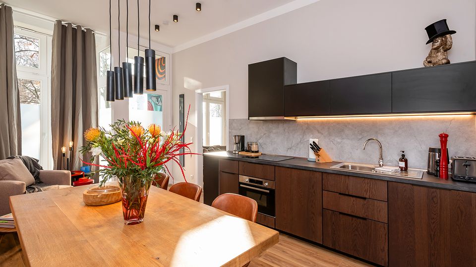 Offene, moderne Wohnküche mit langem Holztisch und hängenden schwarzen Pendelleuchten, daneben eine Küchenzeile mit dunklen Holzfronten, schwarzen Oberschränken und grauer Rückwand mit Unterbauleiste. Große Fenster mit grauen Vorhängen und Holzfußboden sorgen für viel Tageslicht im Raum.