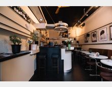 Kleines Café oder Bistro mit moderner Einrichtung. Eine zentrale Theke ist mit Barhockern umgeben, daneben stehen Tische mit braunen Stühlen. An den Wänden hängen Schwarz-Weiß-Fotografien, und Pflanzen sowie Blumen dekorieren den Raum.