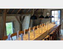 Ein langer, gedeckter Esstisch mit weißen Stoffservietten, Weingläsern und Besteck steht in einem rustikalen Raum mit Holzbalken, Steinwänden und großen Fenstern. Mehrere schwarze Hängeleuchten beleuchten den Tisch.