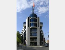 Modernes Bürogebäude mit heller Fassadenverkleidung und großen, dunklen Glasfronten; das Gebäude hat einen symmetrischen Aufbau mit einem zentralen, runden Glasbereich, an dem eine Metallspitze mit roter Fahne angebracht ist. Es befindet sich an einer Straßenecke, umgeben von Gehwegen, einer Ampel und einzelnen Bäumen.