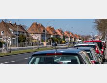 Straßenszene mit Reihenhäusern auf der linken Seite, die rote Ziegeldächer und Gärten haben, sowie einer einspurig verlaufenden Fahrbahn mit parkenden Autos auf der rechten Seite. Im Hintergrund sind Straßenlaternen und ein Schornstein zu sehen, die in eine Vorstadtumgebung eingebettet sind.