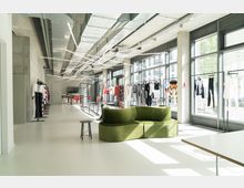 Moderner Ausstellungsraum mit großen Glasfenstern, die Tageslicht hereinlassen. Kleidungsstücke hängen an schlichten schwarzen Kleiderständern, ein grünes Sofa steht im Vordergrund, während im Hintergrund ein Besprechungstisch und dekorative Deckenleuchten zu sehen sind.