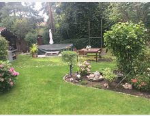 Gepflegter Garten mit einer Rasenfläche, Blumenbeeten, Sträuchern und dekorativen Steinen. Im Hintergrund stehen ein überdachter Pavillon, eine mit Plane abgedeckte Terrasse und ein Holztisch mit Stühlen, umgeben von Bäumen und Hecken.