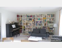 Wohnzimmer mit einer großen, weißen Bücherwand, die mit zahlreichen Büchern und dekorativen Objekten gefüllt ist. Links steht ein schwarzes Klavier mit Notenblättern darauf, davor ein graues Sofa und ein weißer Couchtisch, daneben eine Stehlampe.