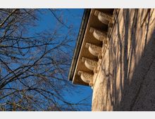 Nahaufnahme einer Gebäudeecke mit verputzter Fassade im Sonnenlicht, versehen mit dekorativen Konsolen unter dem Dachüberstand. Im Hintergrund sind kahle Äste eines Baumes vor klarem, blauem Himmel zu sehen.