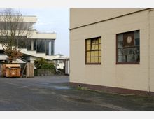 Hinterhof mit gepflasterter Fläche, linkerhand ein modernes weißes Gebäude mit großen Glasfassaden und flachem Dach, rechts eine beige Backsteinwand mit großen Fenstern und teilweise kaputten Scheiben. Im Hof stehen ein Müllcontainer, ein Holzzaun und ein Fahrrad.