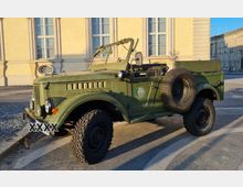 Ein olivgrüner Geländewagen im Retro-Stil mit offenem Verdeck und Reserverad an der Seite, geparkt vor einer cremefarbenen Gebäudefassade mit großen Fenstern.