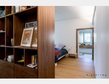 Ein moderner Innenraum mit einem Schlafzimmer und einem angrenzenden Badezimmer. Links im Vordergrund steht ein offenes Holzregal mit Büchern, Bilderrahmen und Dekoration, während das Schlafzimmer ein Bett mit bunter Bettwäsche und Holzboden zeigt; im Hintergrund ist ein weiß gefliestes Badezimmer mit Waschbecken und Fenster sichtbar.