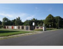 Eine Reihenanlage aus einfachen Garagen mit verschiedenfarbigen Türen, umgeben von Rasenflächen und Bäumen. Im Vordergrund verläuft eine asphaltierte Straße, daneben befindet sich ein Bürgersteig und ein kleines Schild mit einer Aufschrift.
