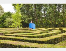 Labyrinth aus niedrigen, gepflegten Hecken in einem Park, umgeben von hohen Bäumen; in der Mitte befindet sich eine blaue Rutsche mit einer kleinen Überdachung.